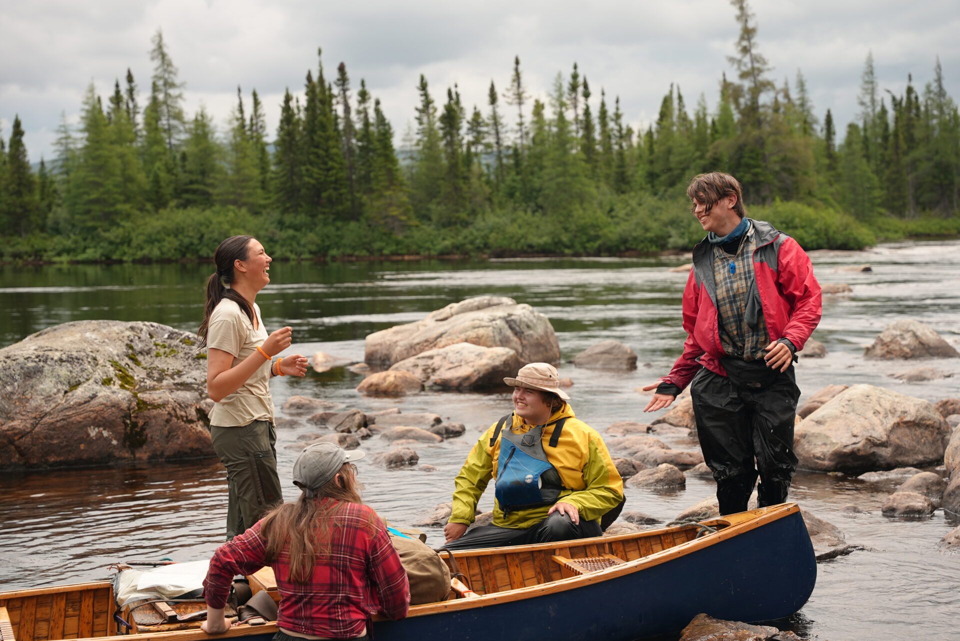 Friends laughing by canoe in scenic riverside.