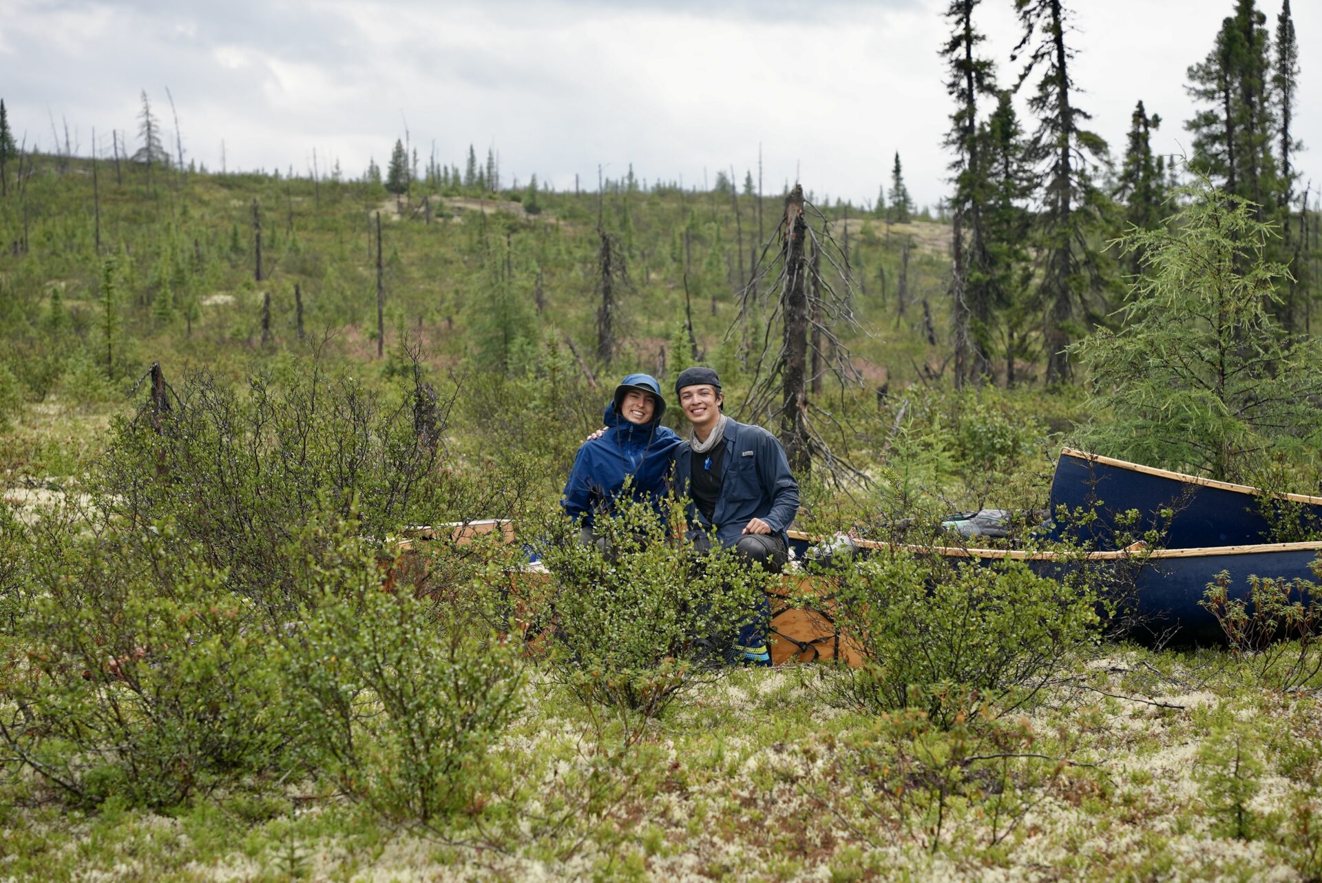 Two people sitting near canoes in a forest.