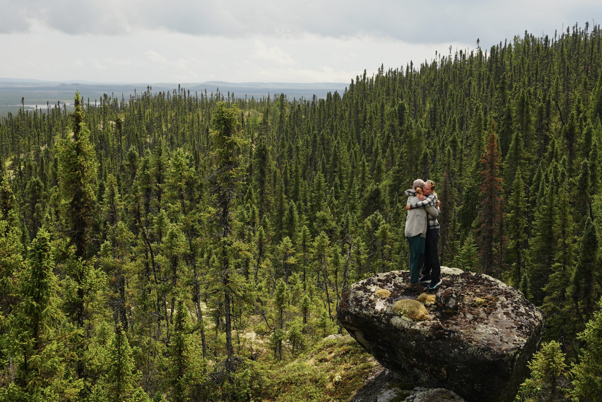 Couple hugging atop large rock in forest.