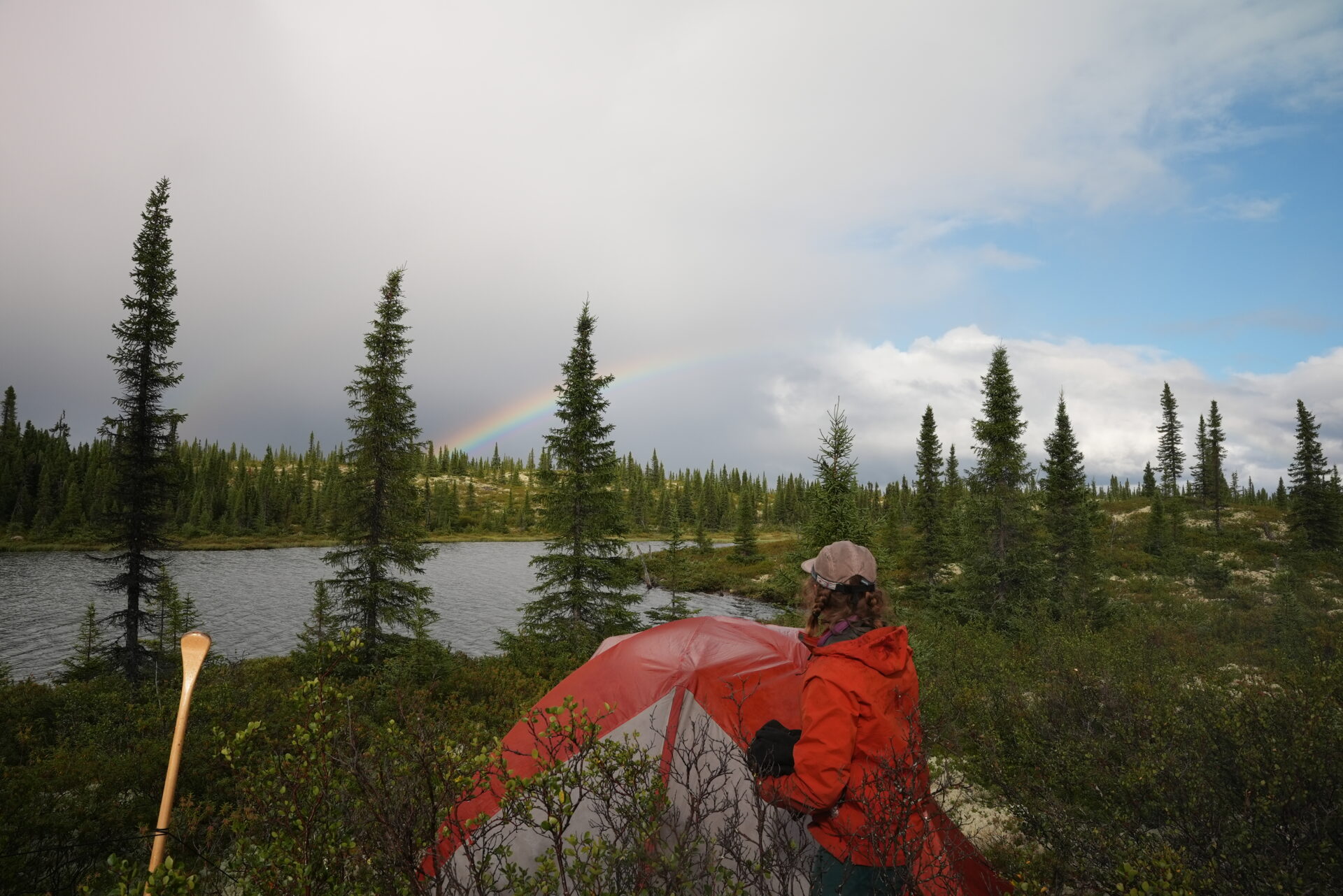 Camper by tent, rainbow over forest lake