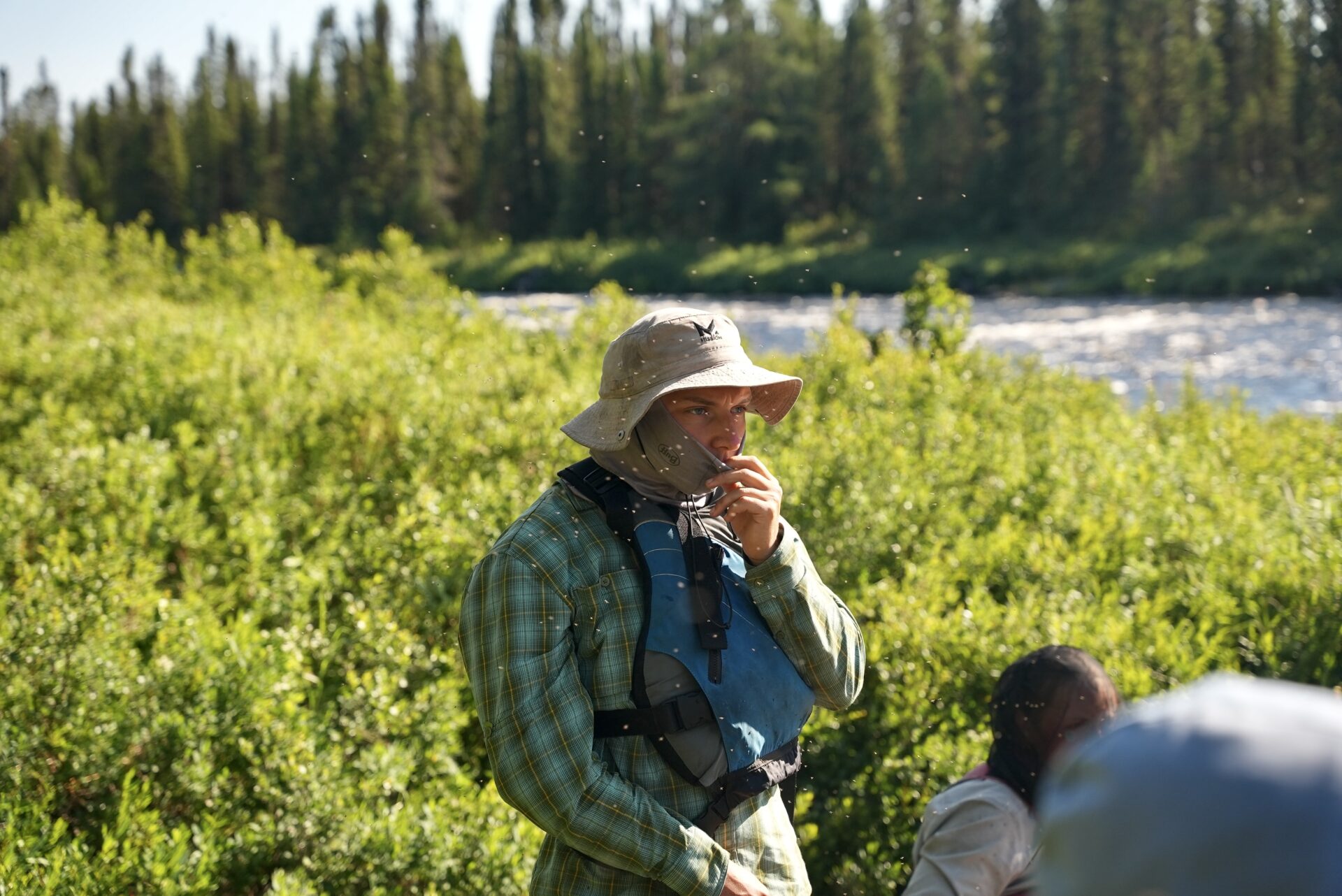 Person outdoors by river wearing hat and face covering.