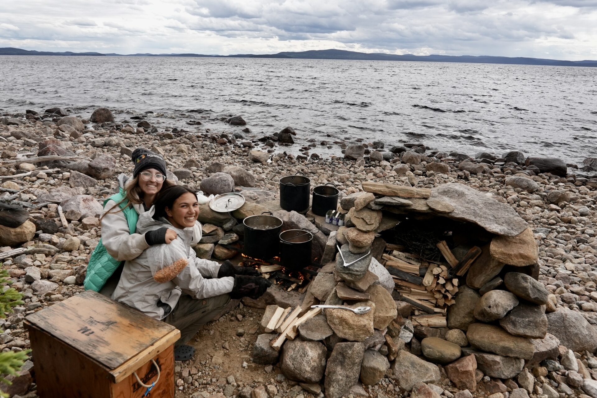 Two people cooking by lakeside campfire.