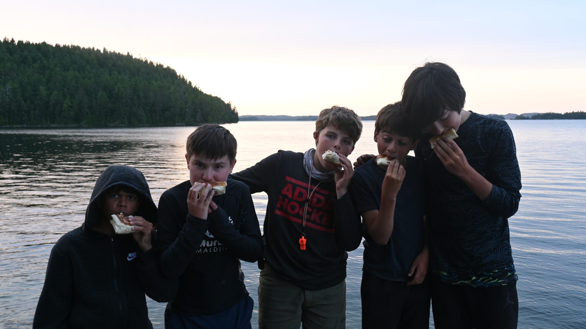 Kids eating sandwiches by a lake at sunset.