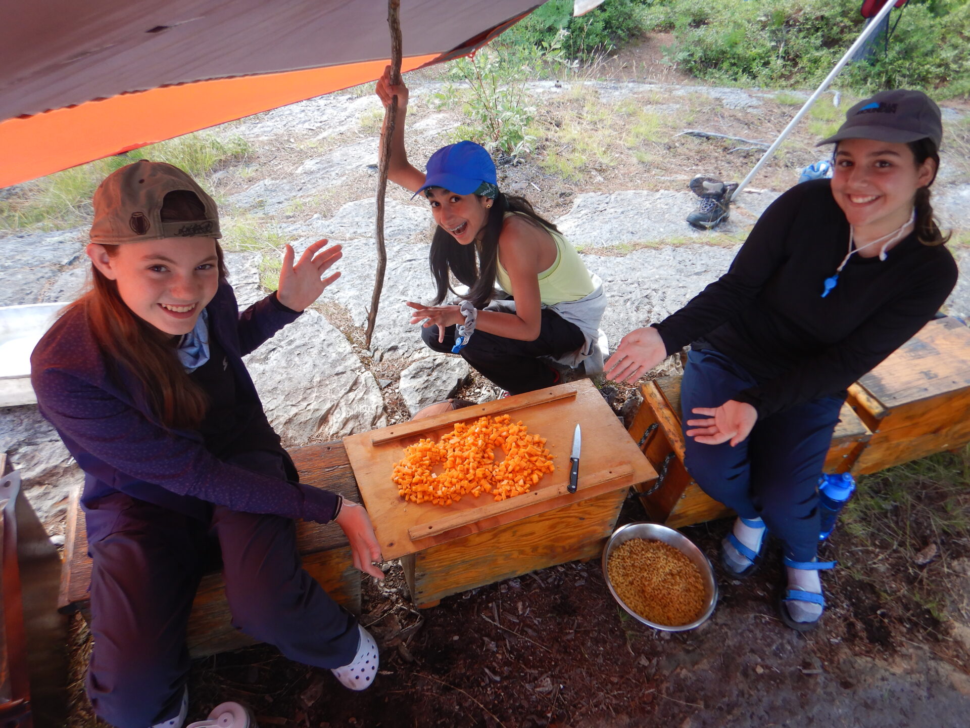 Three girls chopping vegetables at outdoor campsite.