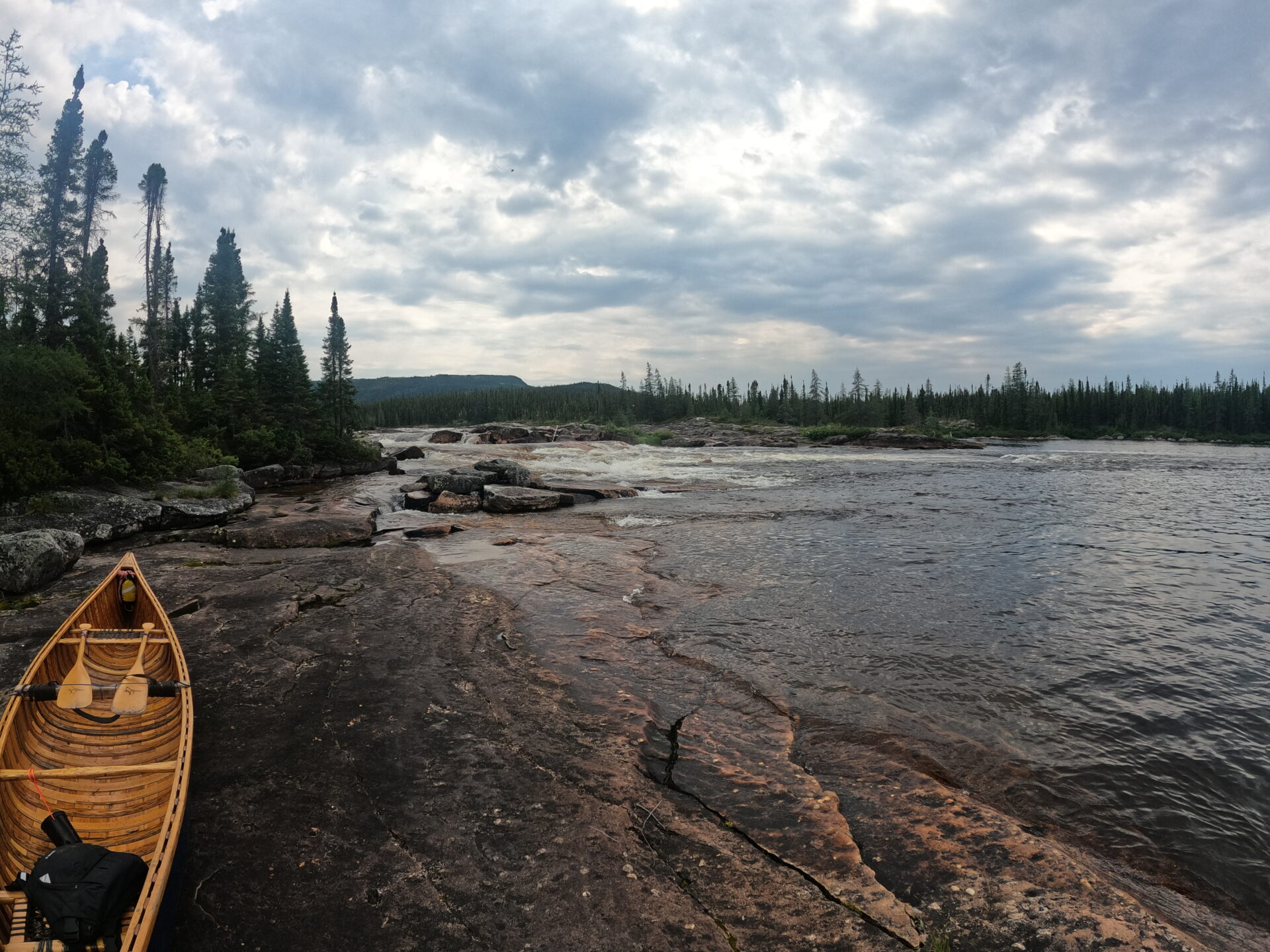 Wooden canoe by rocky riverbank forest and cloudy sky.