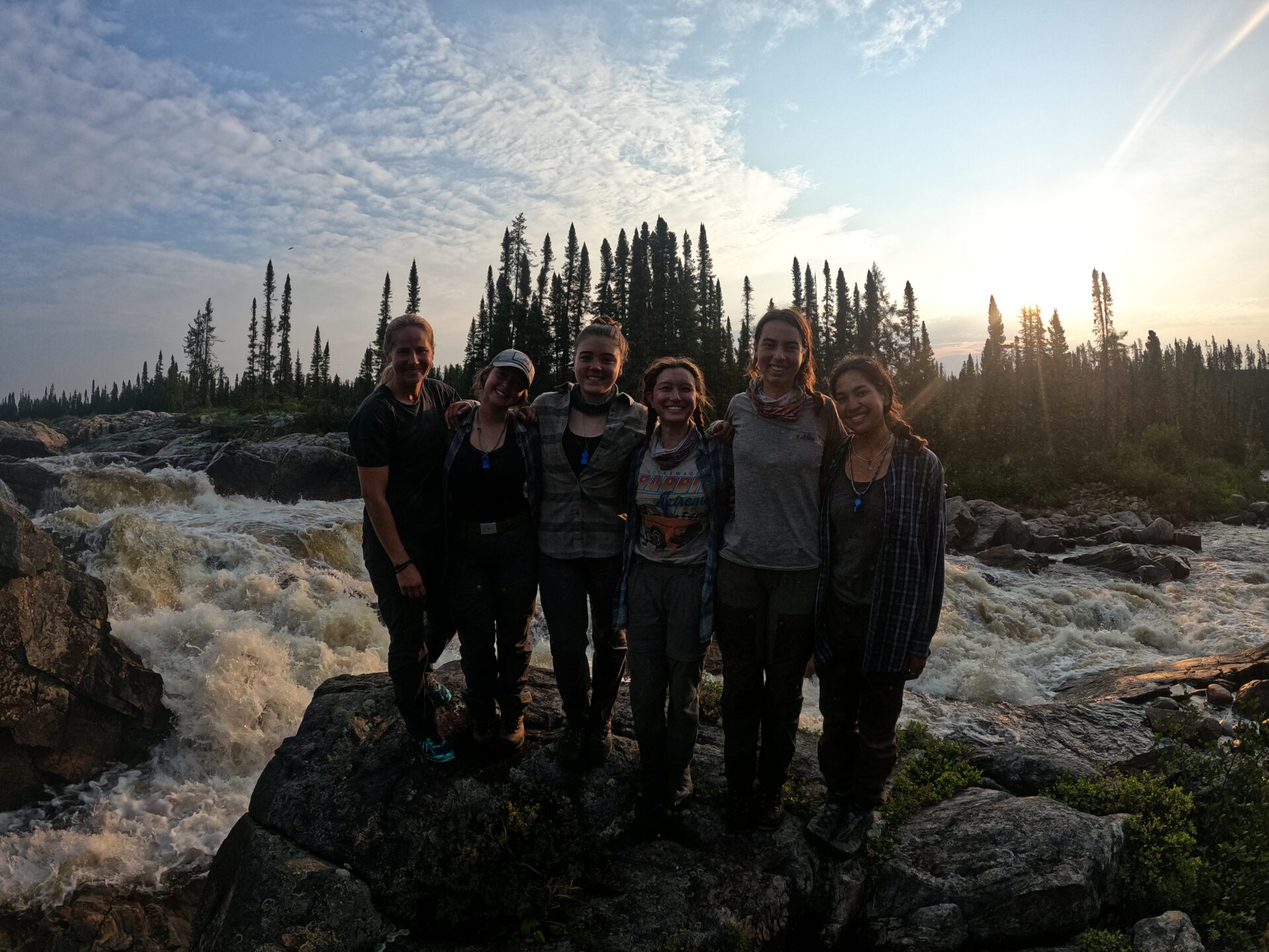 Group posing by river in forest, sunset background.