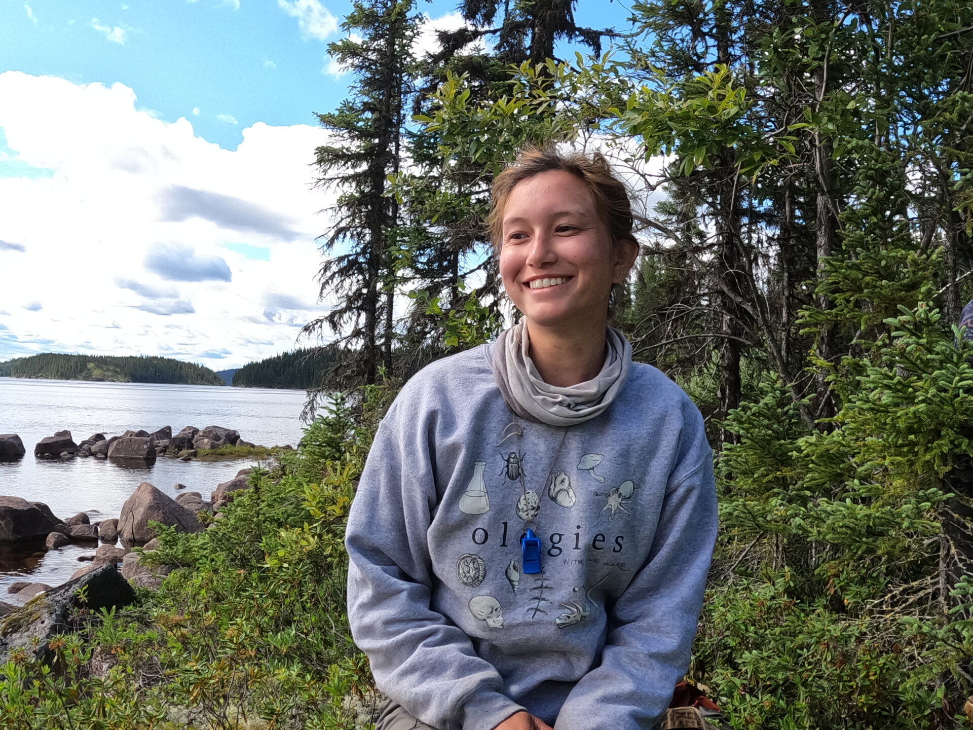 Woman smiling outdoors by a lake and forest.