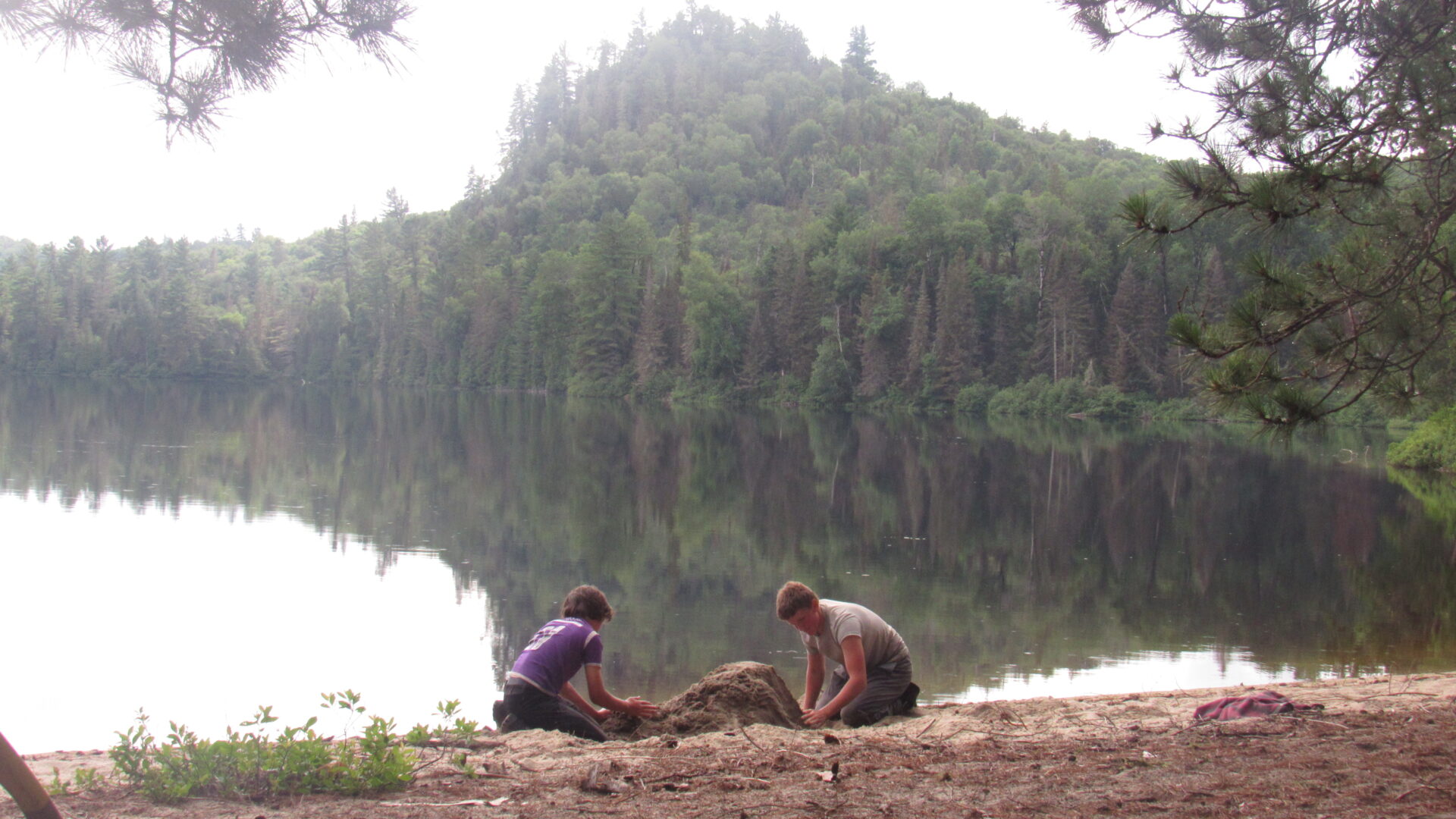 Two boys building sandcastle by lake with forest hills.