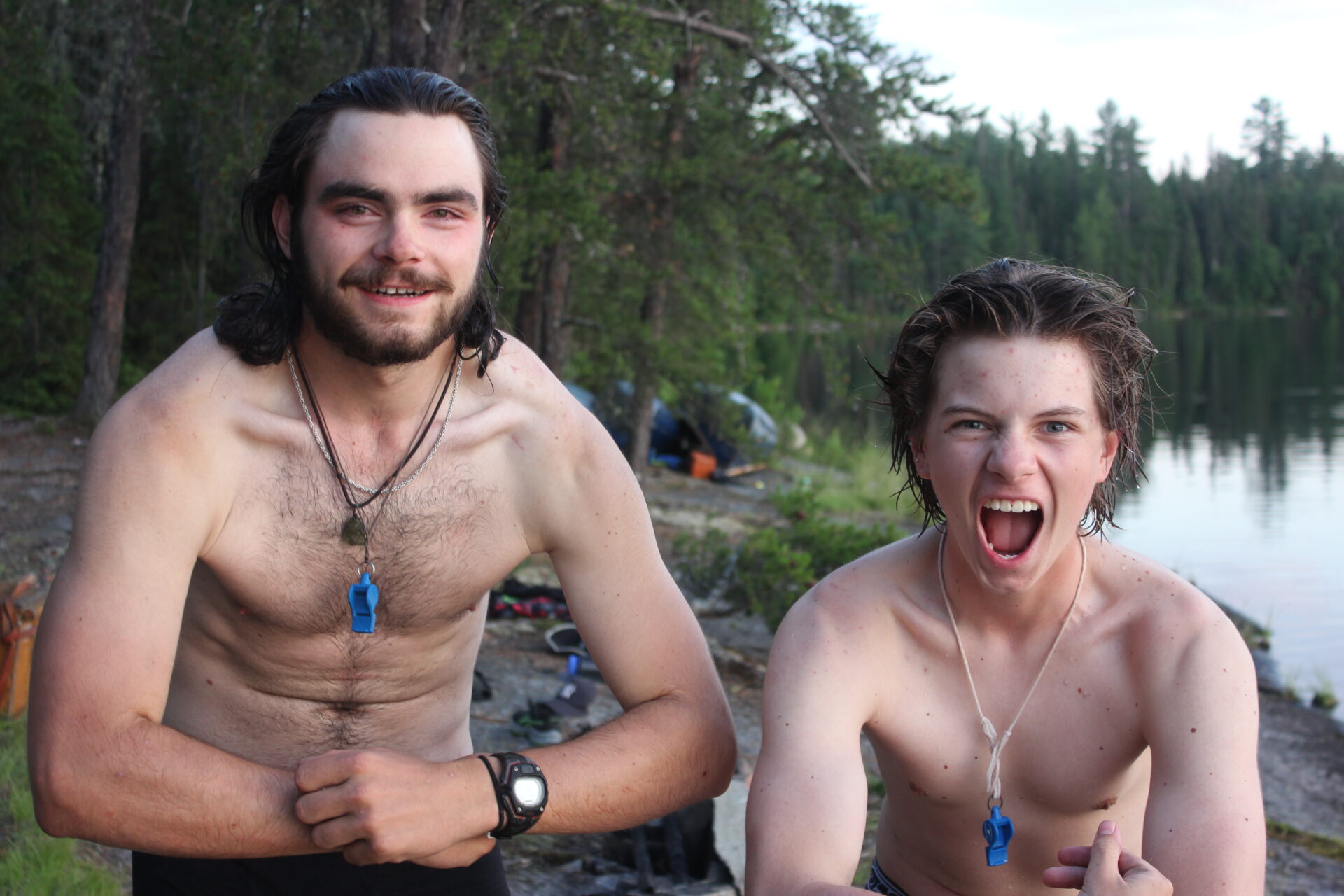 Two shirtless men smiling near a forest lake.