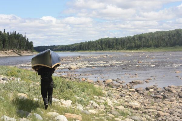 Person portaging canoe beside river and forest.