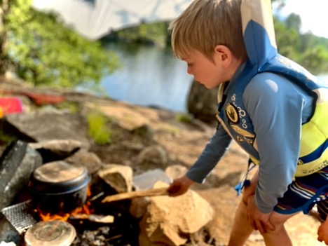 Child cooking over campfire by a lake.